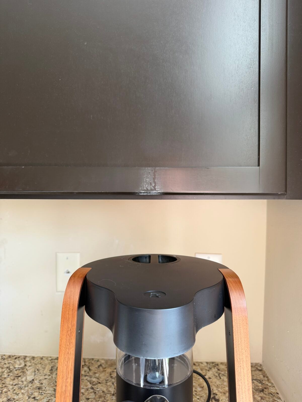 A black and wood coffee maker sits on a speckled granite countertop beneath a dark brown kitchen cabinet, with electrical outlets visible on the beige wall in the background.