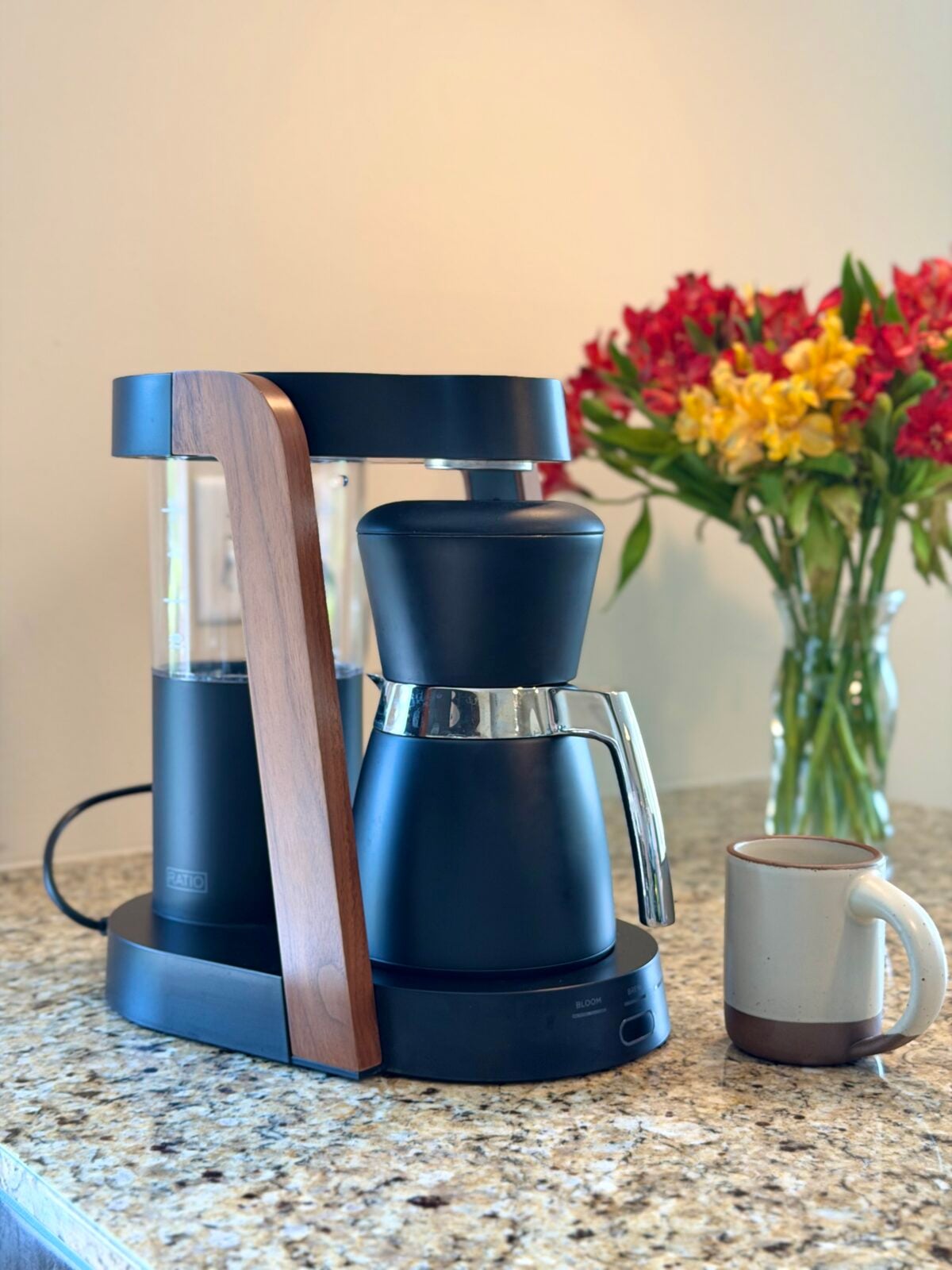 A black and silver coffee maker with a wooden accent sits on a granite countertop next to a beige mug. A clear vase filled with red and yellow flowers is in the background.