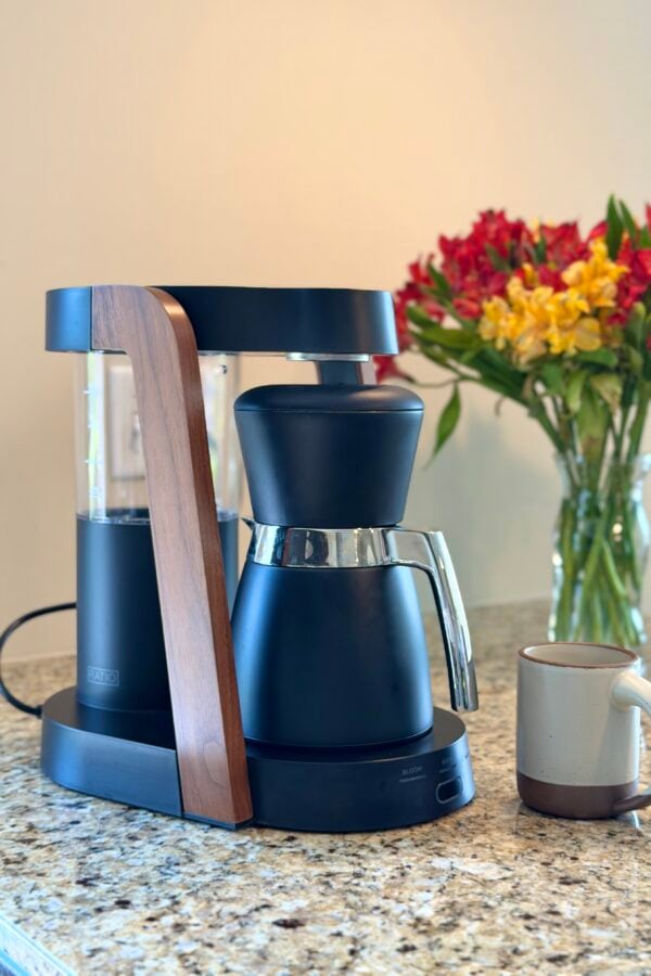 A black and silver coffee maker with a wooden accent sits on a granite countertop next to a beige mug. A clear vase filled with red and yellow flowers is in the background.