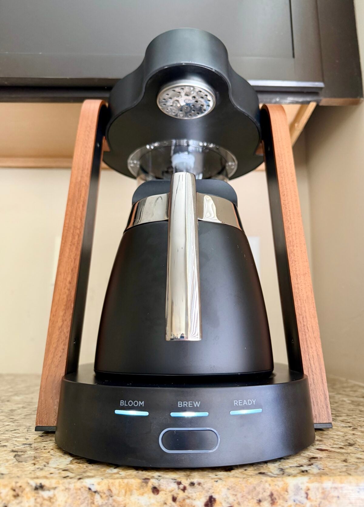 A black and silver coffee maker with wooden side handles sits on a granite countertop. The base shows three labeled buttons: Bloom, Brew, and Ready. The coffee maker is positioned under a cabinet.