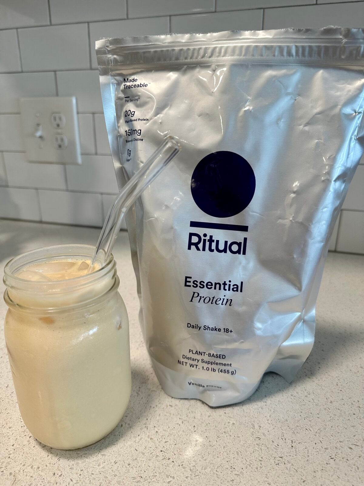 A glass jar with a creamy drink and a glass straw sits next to a large silver bag labeled Ritual Essential Protein on a kitchen countertop. White subway tile backsplash and electrical outlets are visible in the background.