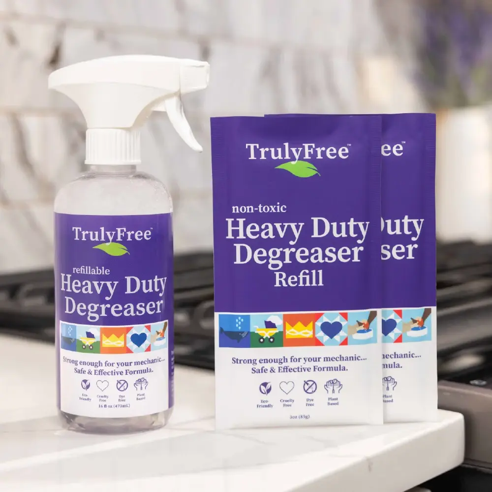 A spray bottle and two refill packets of Truly Free Heavy Duty Degreaser are placed on a kitchen counter near a stove. The labels highlight that the product is non-toxic and safe for everyday use.