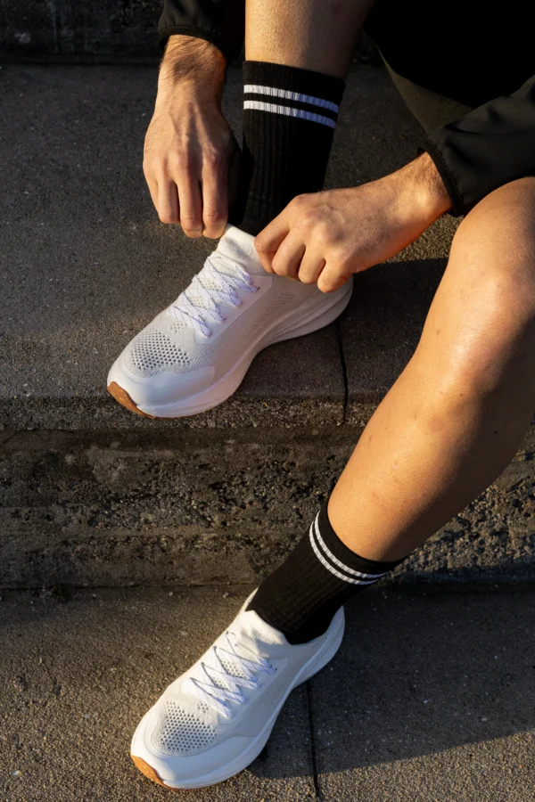 A person sitting on outdoor concrete steps ties the laces on a white athletic shoe, wearing black shorts and black socks with two white stripes.