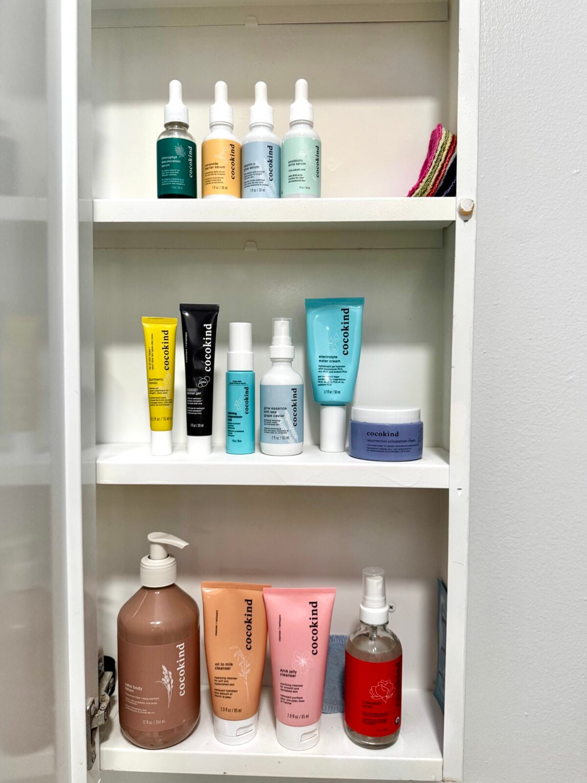 Three white shelves inside a cabinet hold various skincare and personal care products, including bottles, tubes, and jars in different colors, with washcloths on the top right shelf. The cabinet is against a white wall.