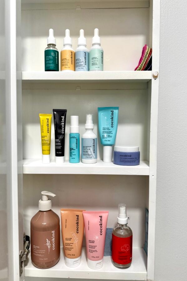 Three white shelves inside a cabinet hold various skincare and personal care products, including bottles, tubes, and jars in different colors, with washcloths on the top right shelf. The cabinet is against a white wall.