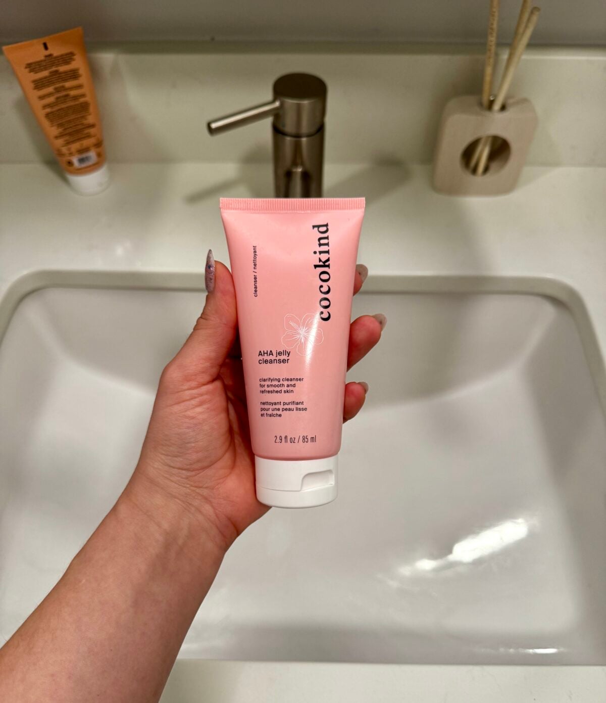 A hand holding a pink tube of Cocokind AHA Jelly Cleanser over a white bathroom sink, with a soap dispenser and a container holding sticks visible on the countertop.