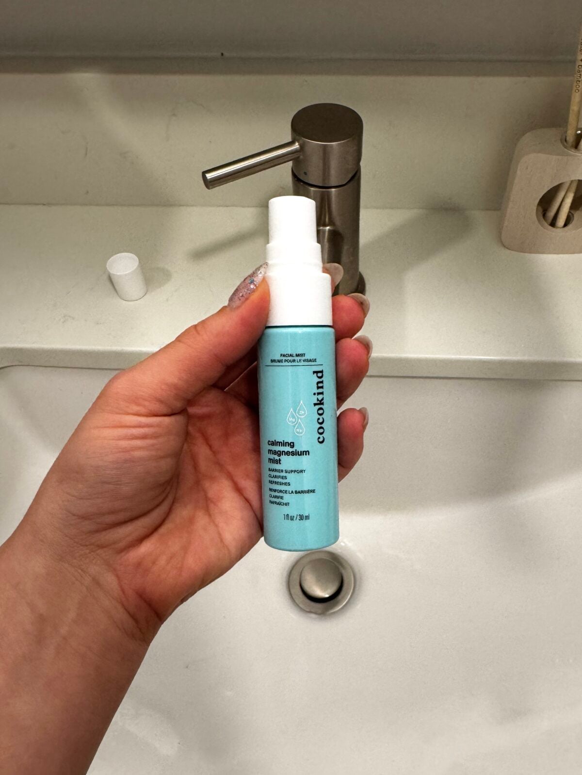 A person holds a blue spray bottle labeled Calming Magnesium Mist from the brand cocokind over a white sink with a silver faucet. The bottle cap is placed on the counter.