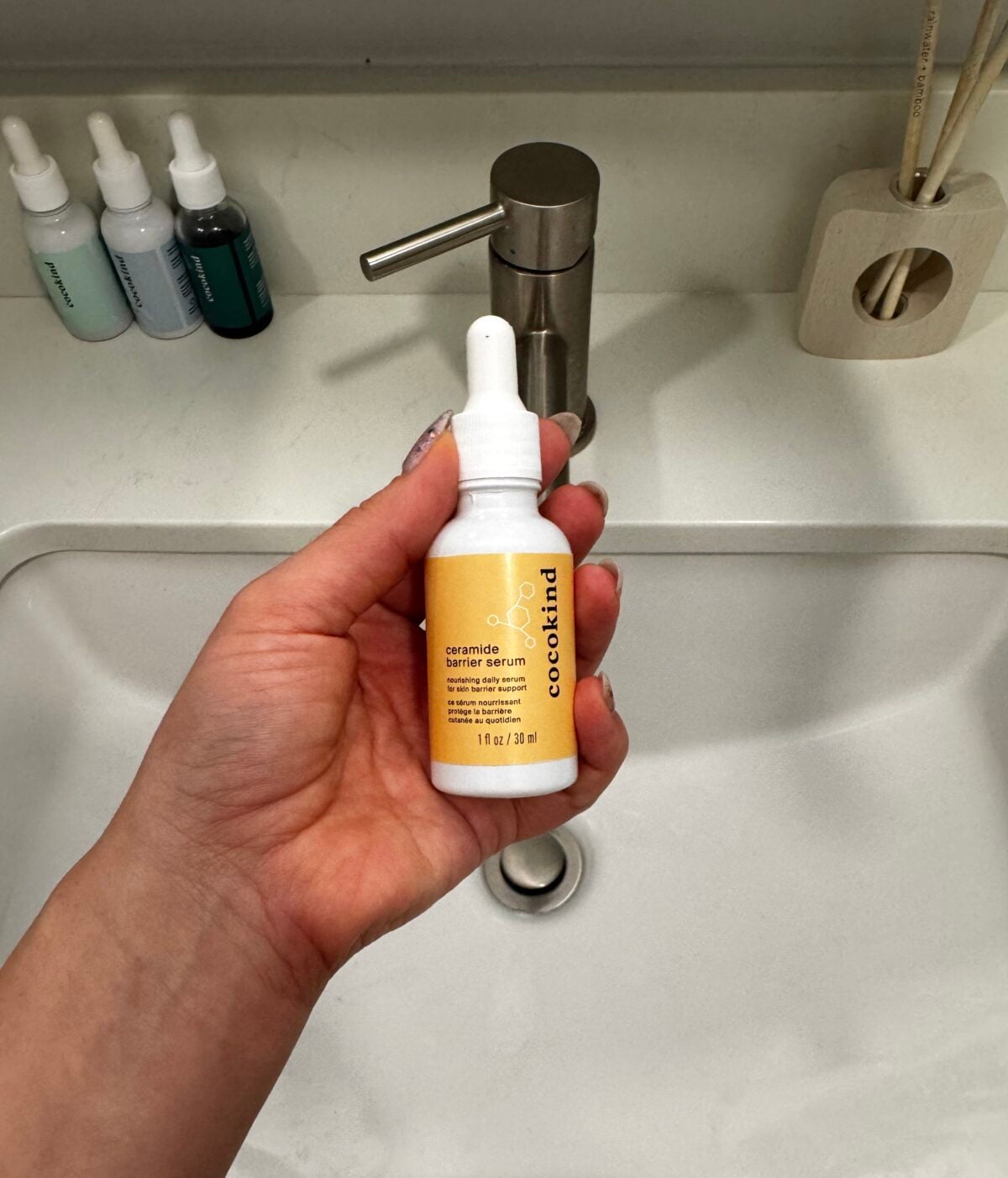 A hand holds a bottle of Cocokind Ceramide Barrier Serum with a dropper in front of a white bathroom sink and faucet; other skincare bottles are on the countertop in the background.