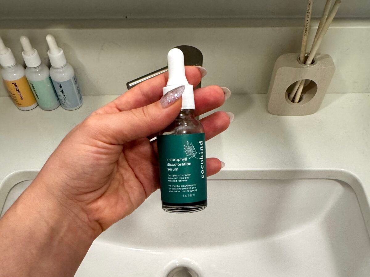 A hand with manicured nails holds a bottle of Good Molecules chlorophyll discoloration serum above a white bathroom sink. Skincare bottles and diffuser sticks are visible on the counter in the background.