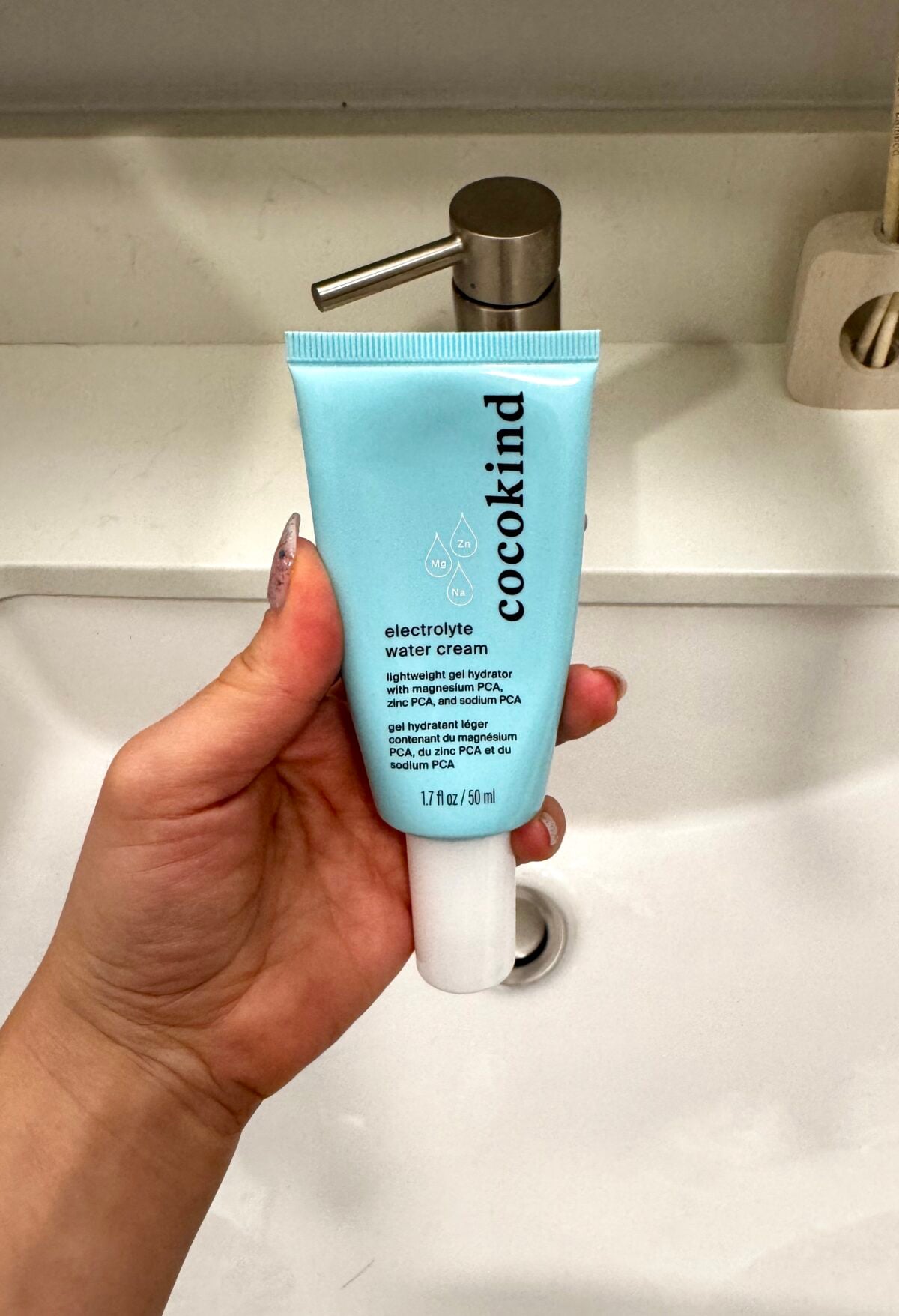 A hand holds a blue tube of Cocokind electrolyte water cream in front of a bathroom sink with a silver pump dispenser. The label describes it as a lightweight, hydrating moisturizer in a 50 ml container.