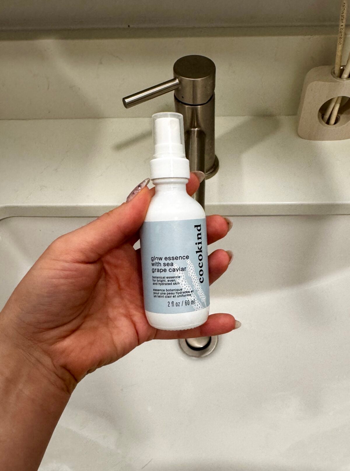 A hand holds a white bottle labeled cocokind glow essence with grape caviar in front of a sink with a silver faucet. The environment appears clean and minimalistic.