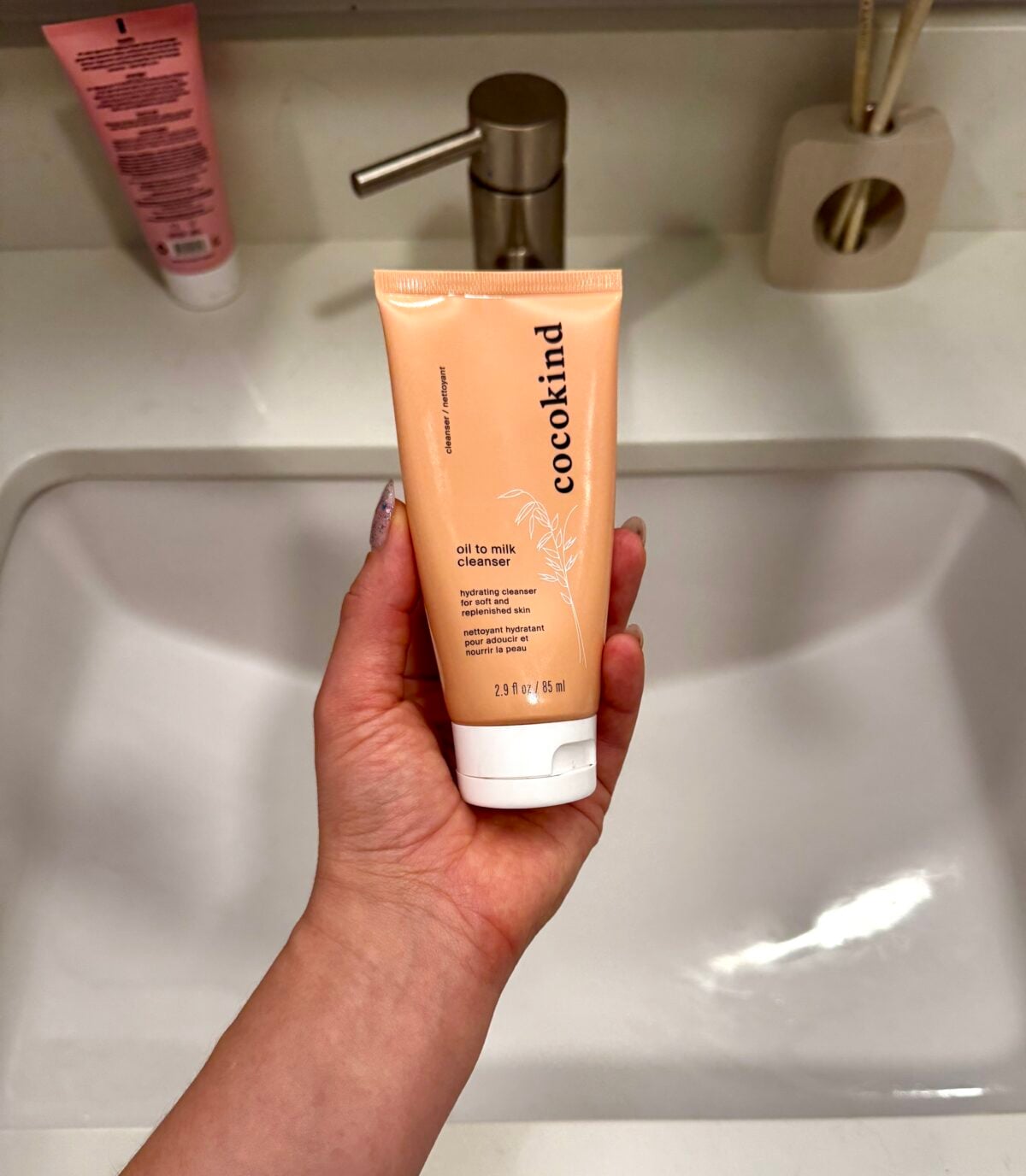 A hand holding a tube of Cocokind Oil to Milk Cleanser over a white bathroom sink; other toiletries and a toothbrush holder are visible on the counter in the background.