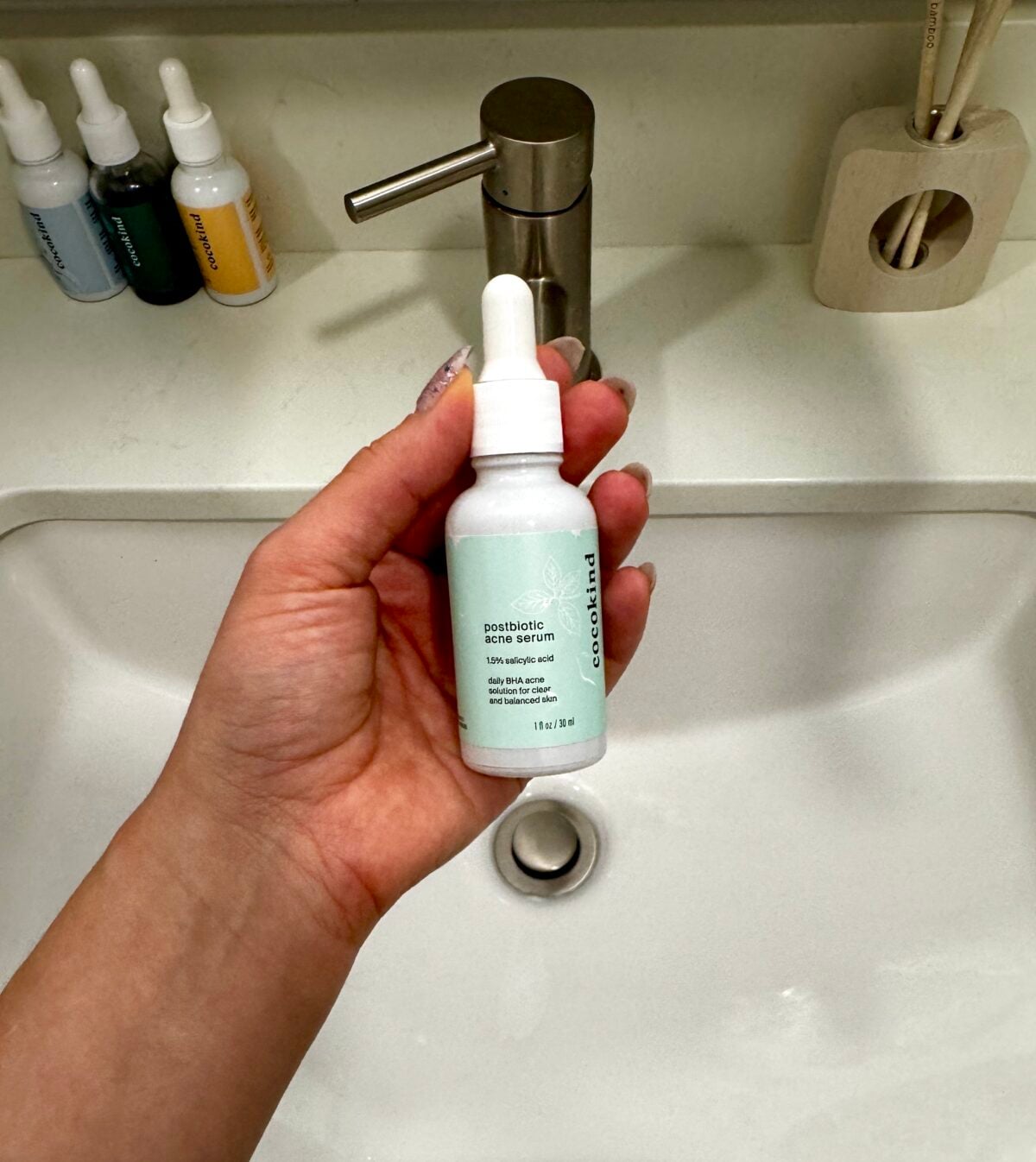 A hand holds a bottle of Cocokind Postbiotic Acne Serum over a white bathroom sink, with three other skincare bottles, a soap dispenser, and a toothbrush holder visible in the background.