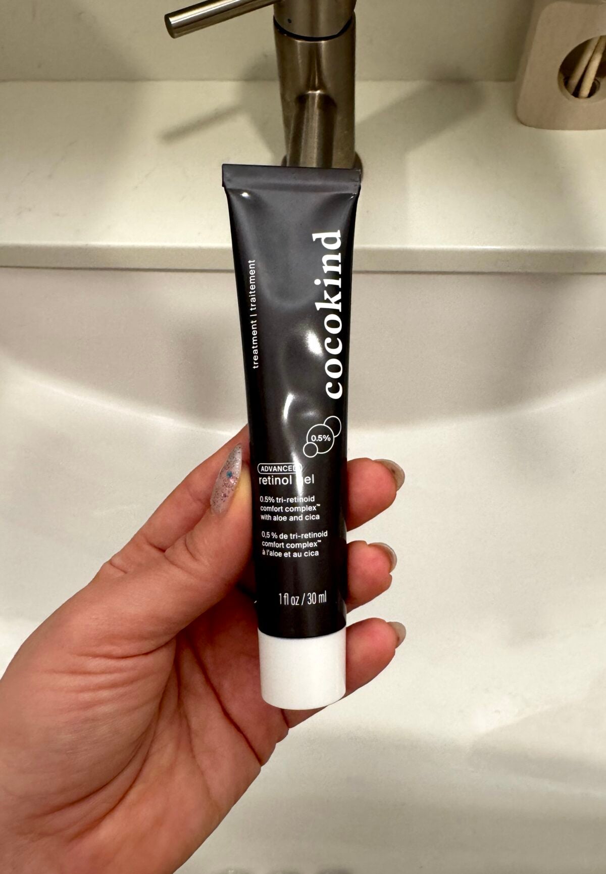 A hand holds a tube of Cocokind 0.05% retinal night cream in front of a white sink with a silver faucet. The black tube displays product details in white text.