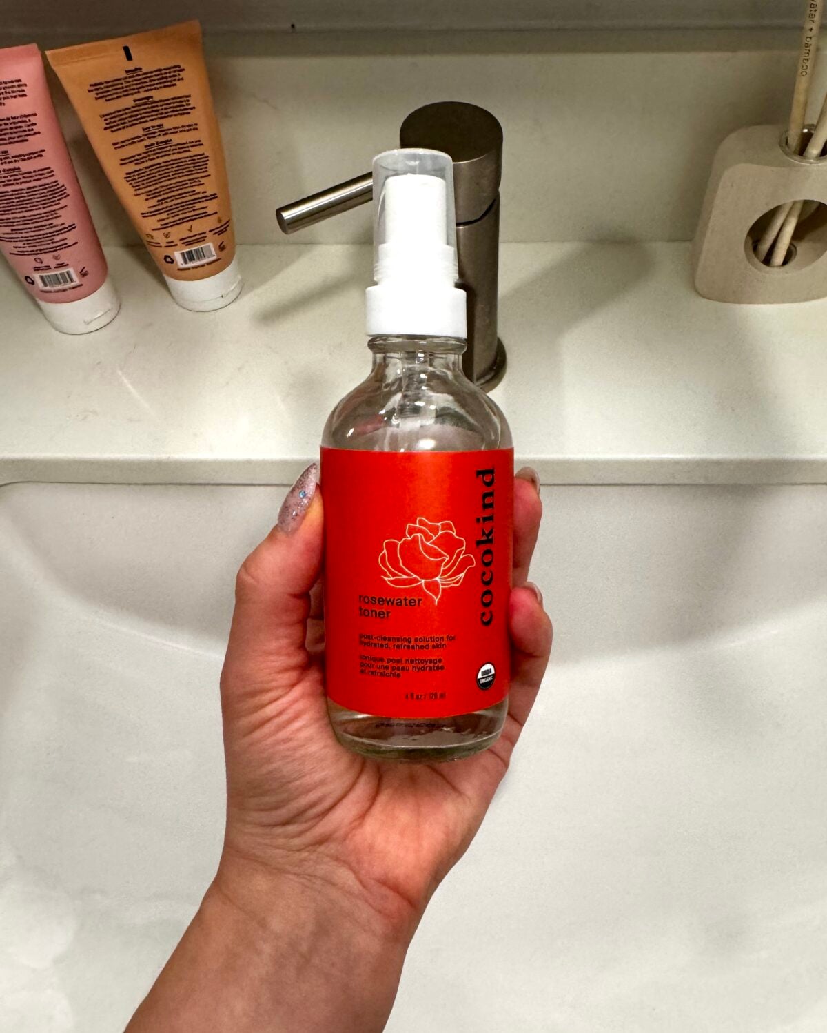 A hand is holding a clear glass spray bottle labeled &ldquo;cocokind rosewater toner&rdquo; in front of a bathroom sink with other skincare products and a faucet visible in the background.