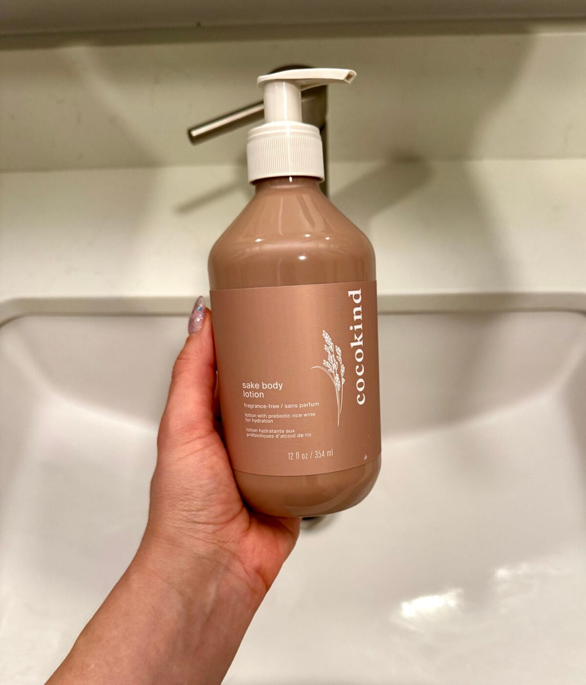 A hand holds a bottle of Cocokind sake body lotion with a pump dispenser, positioned over a white sink. The bottle is beige with white text and a small illustration of rice stalks.