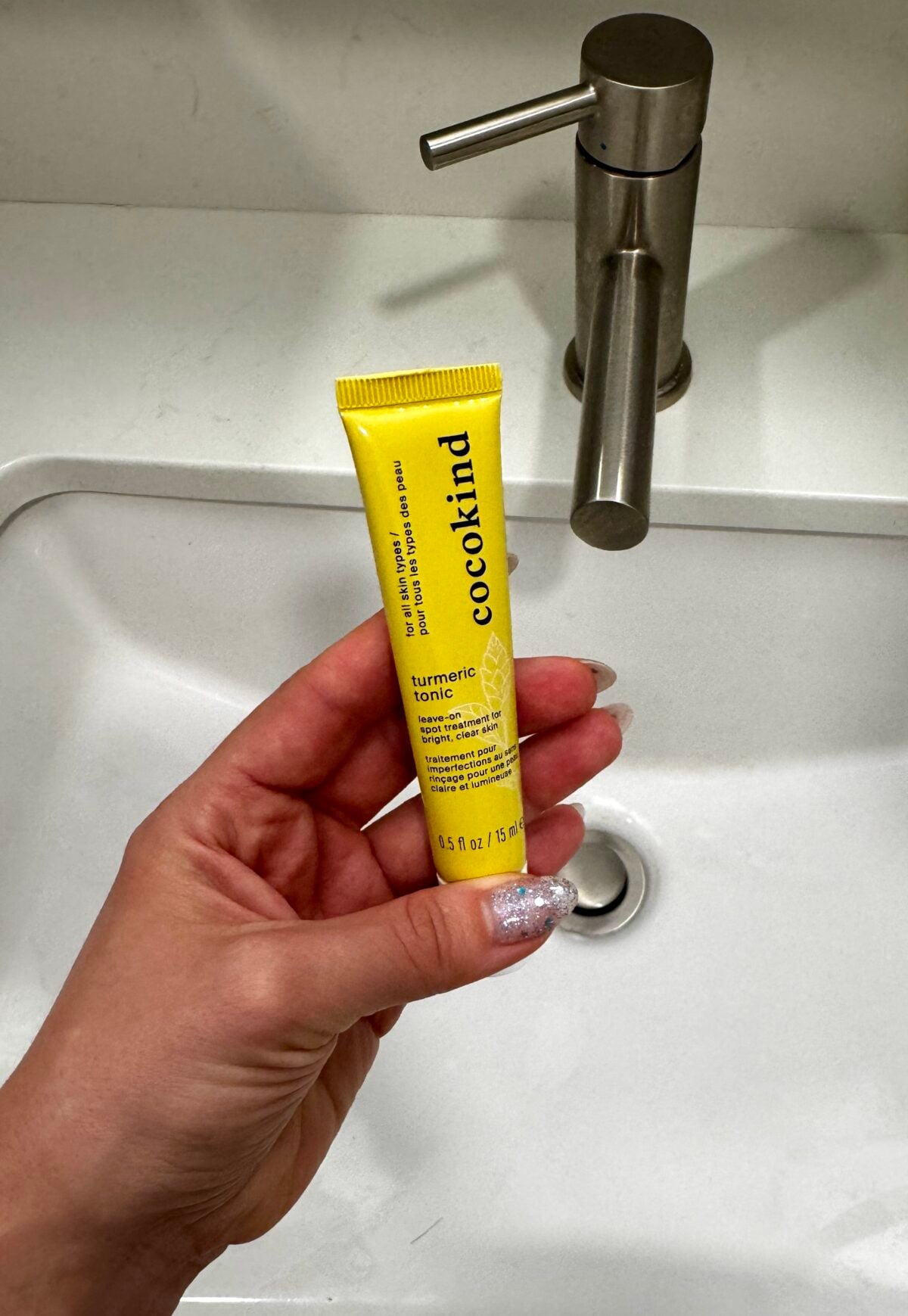 A hand with glittery nails holds a yellow tube of Cocokind Turmeric Tonic above a white sink with a silver faucet.