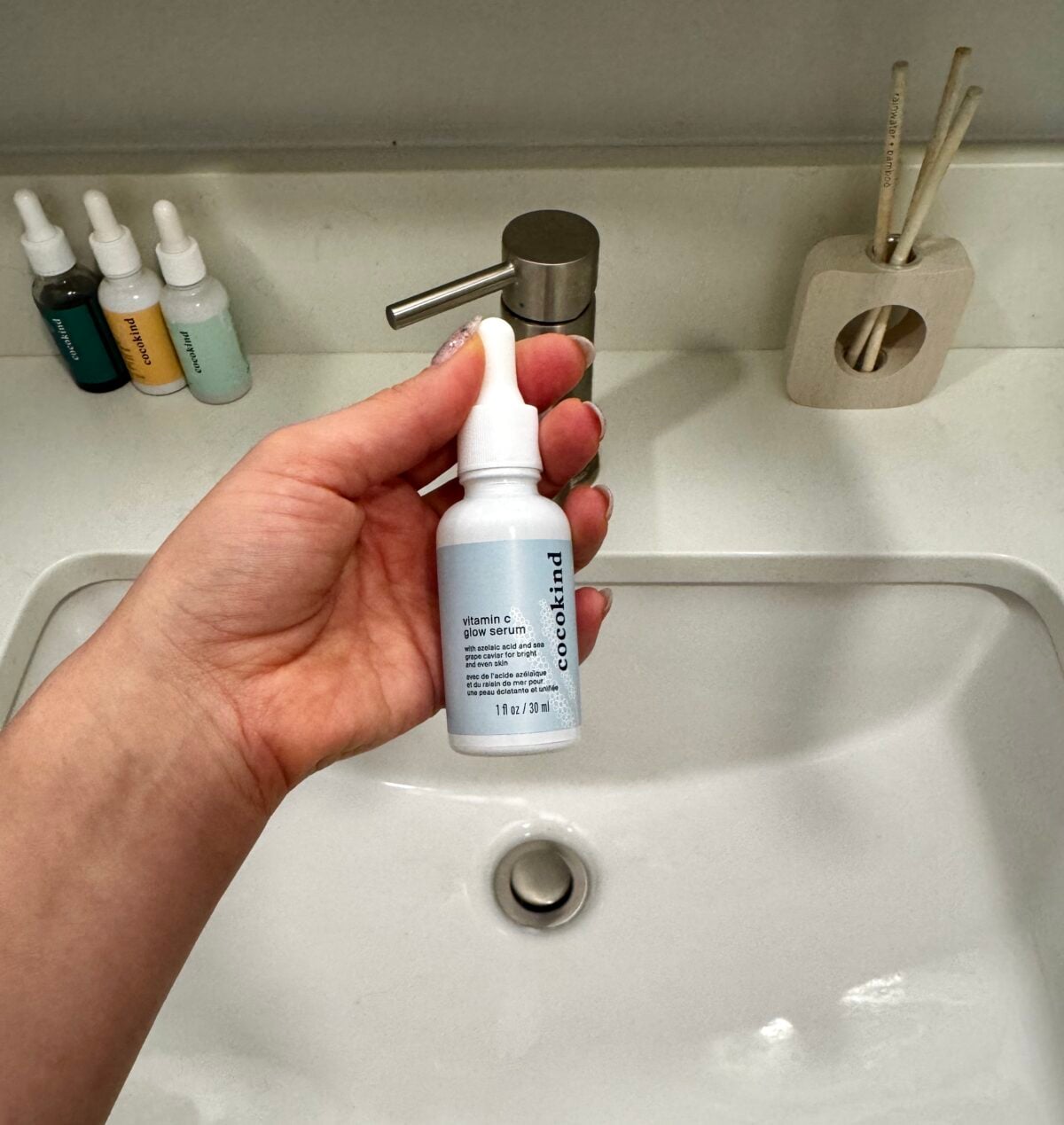A hand holds a dropper bottle labeled Cocokind Vitamin C Glow Serum above a white bathroom sink. Three other dropper bottles and a reed diffuser are on the counter in the background.
