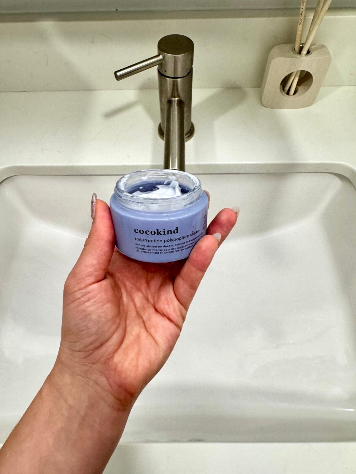 A hand holds an open jar of Cocokind Resurrection Polyphenol Cleanser with visible white cream inside, in front of a bathroom sink and metallic faucet.