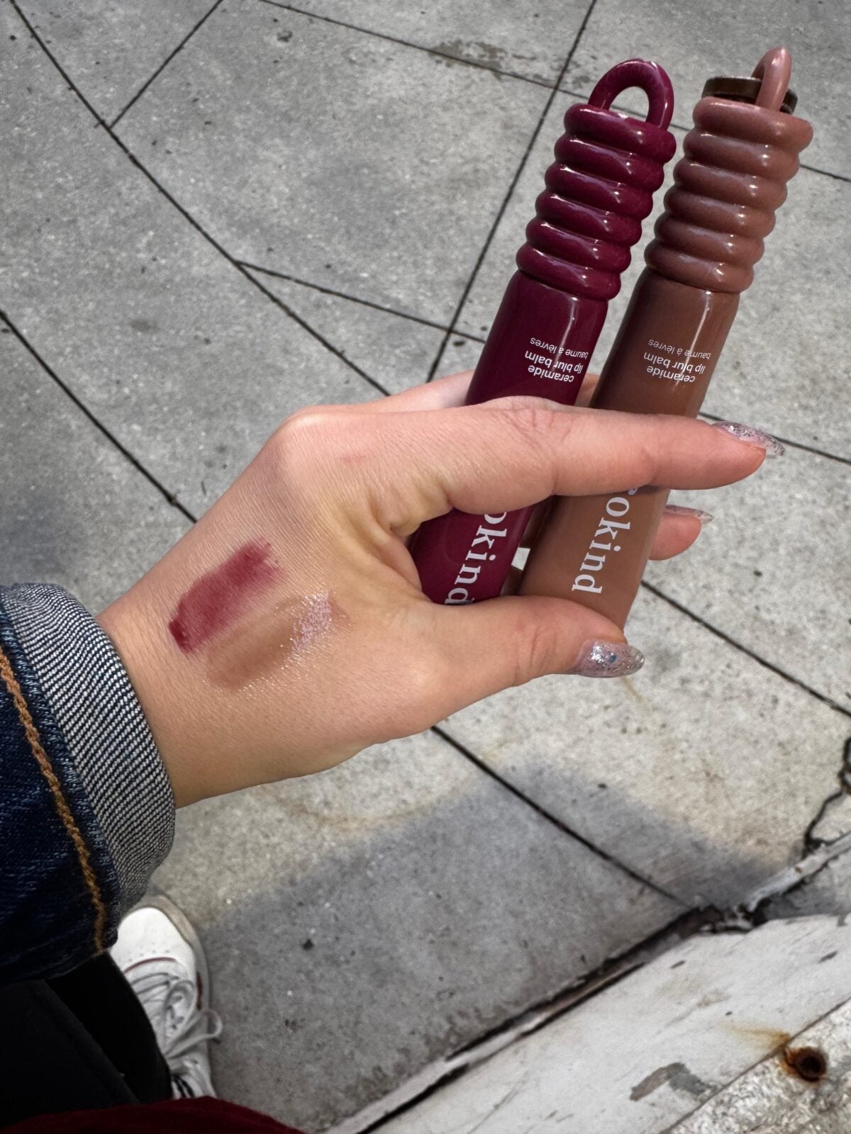 A hand holding two lipstick tubes, one burgundy and one nude. Swatches of both lipstick colors are visible on the back of the hand. The background is a concrete sidewalk.