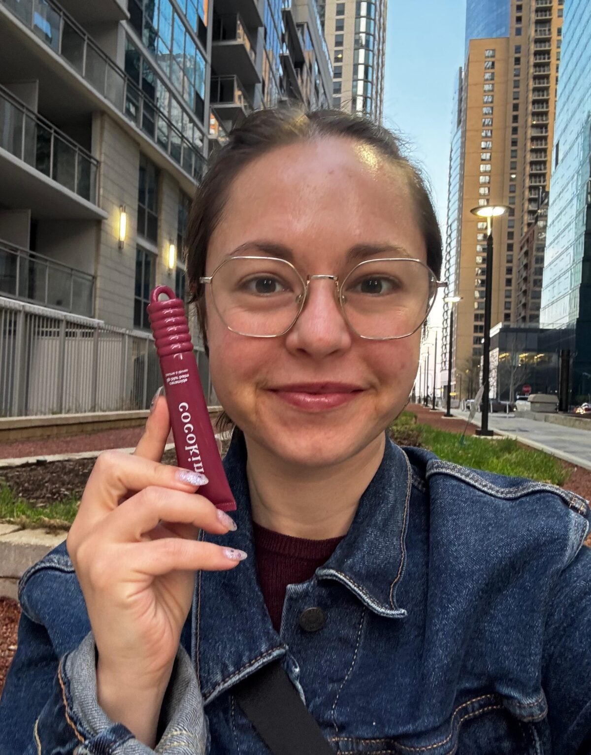 A person with glasses and a denim jacket smiles while holding a purple tube labeled GOODPUP outdoors on a city sidewalk with tall buildings in the background.