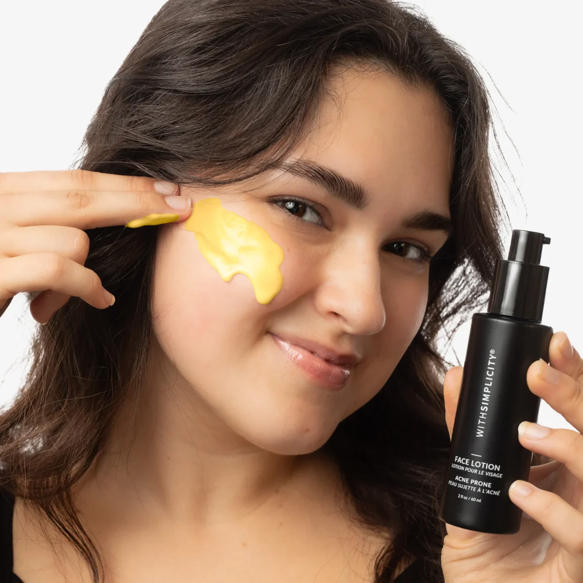 A person smiles while applying a yellow cream to their cheek with their fingers, holding a black bottle labeled Face Lotion in the other hand against a white background.