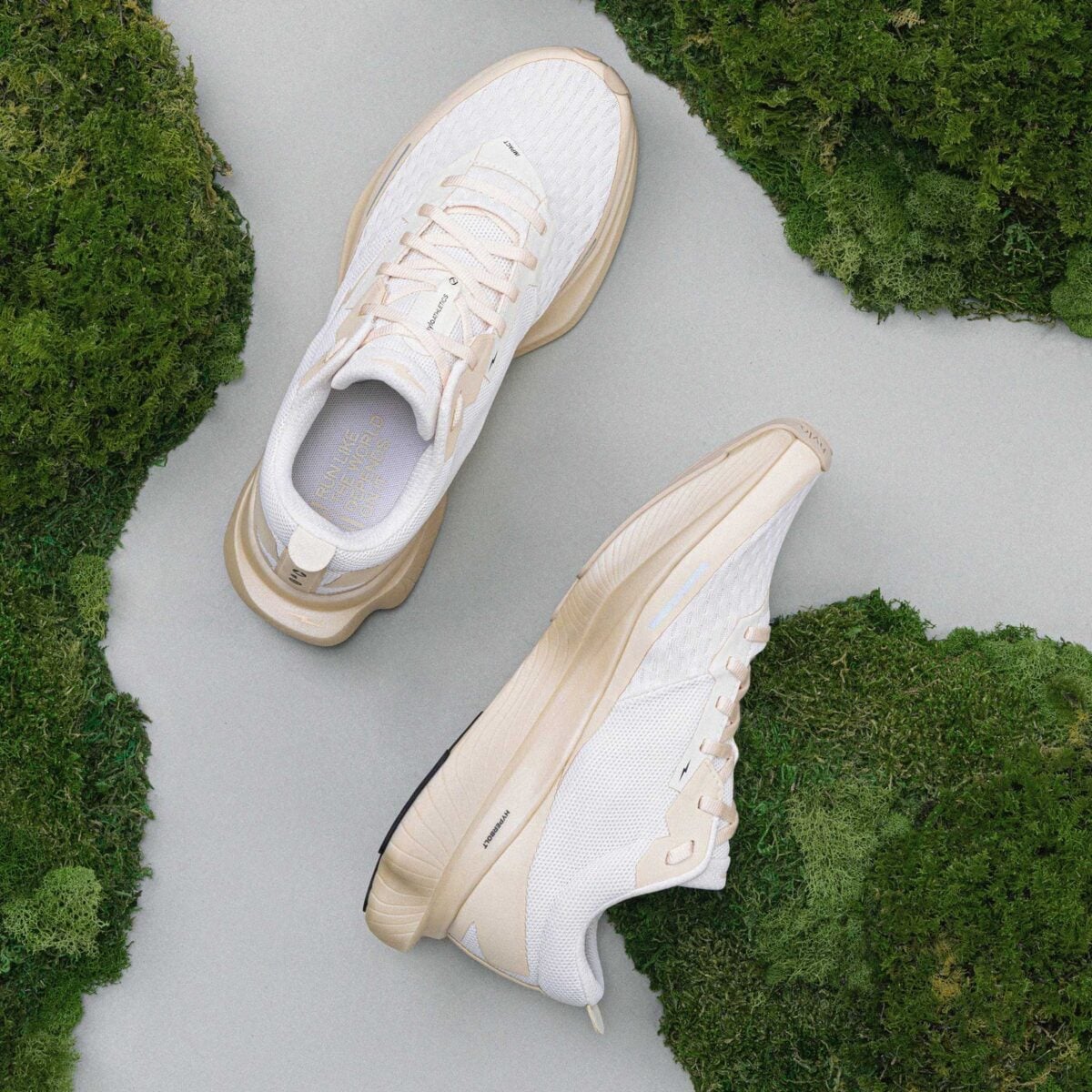 A pair of white and beige athletic shoes are displayed on a light gray surface surrounded by patches of green moss. The left shoe is upright and the right shoe is on its side, showing the sole.