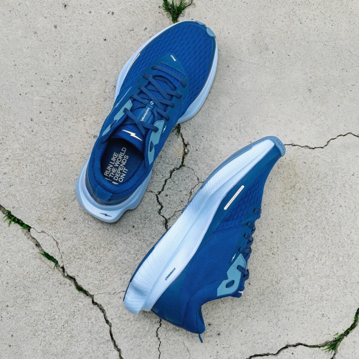 A pair of blue athletic running shoes with white soles are placed on a cracked, light-colored concrete surface with small patches of grass.
