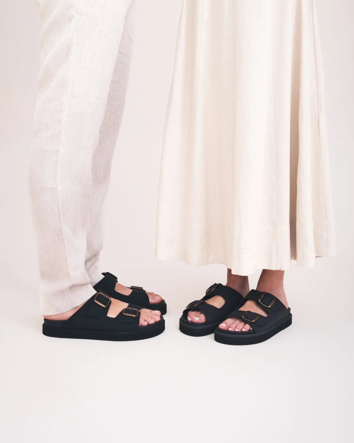 Two people stand facing each other, shown from the knees down. One wears light-colored pants and the other a light, long skirt. Both wear black sandals with buckle straps. The background is plain and light.