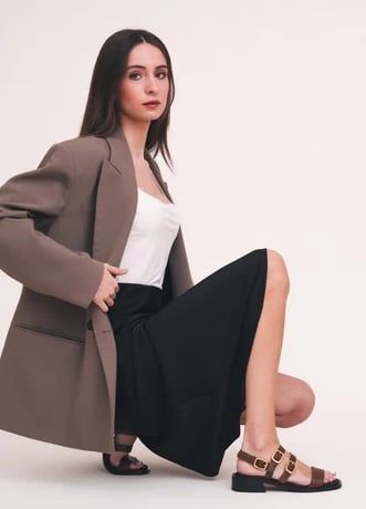 A woman with straight brown hair wears a tan blazer, white top, black skirt, and brown sandals with buckles. She is kneeling on one knee against a plain light background.