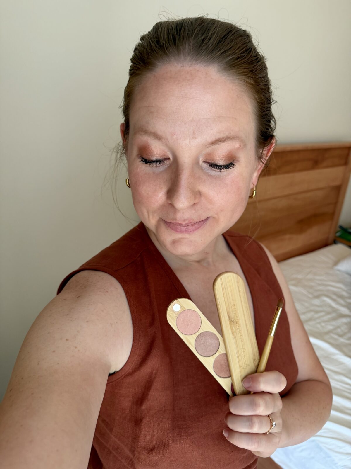 A person with light skin and brown hair pulled back holds an eyeshadow palette and brush while looking down, showing natural makeup. The background includes a wooden headboard and white bedding.