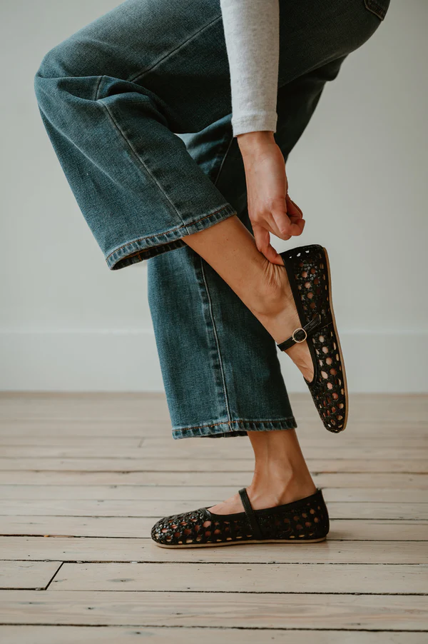 A person wearing blue jeans and a light long-sleeve top bends their leg and touches their ankle while wearing black woven Mary Jane shoes, standing on a light wooden floor.