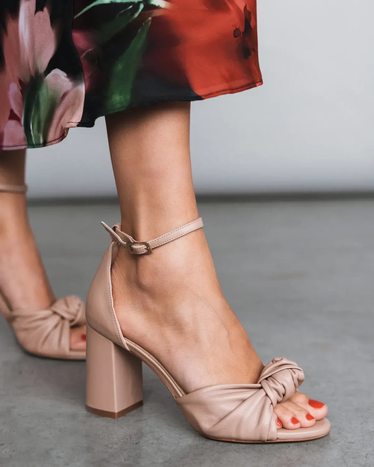 A person wearing open-toe, nude-colored block heel sandals with ankle straps and a knotted design, paired with a floral skirt, standing on a gray floor.