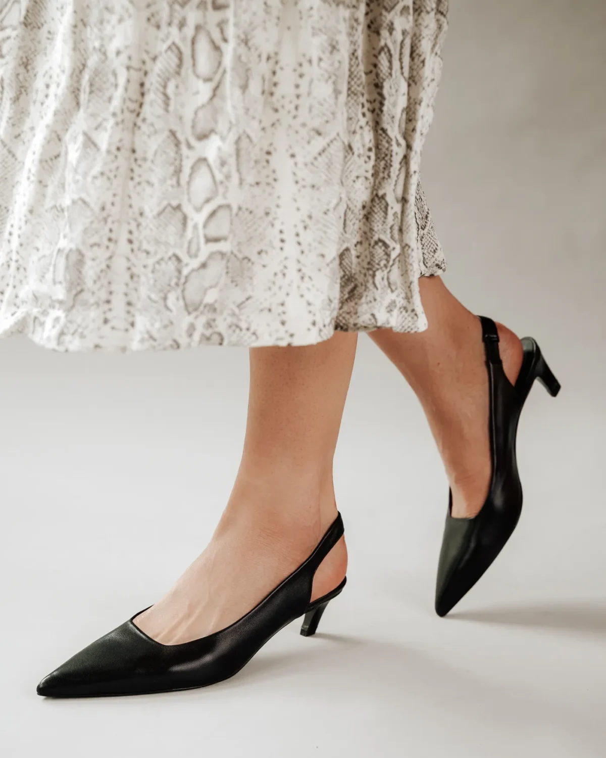 A person wearing a white, snakeskin-patterned skirt and black pointed-toe slingback heels with low kitten heels stands on a light-colored surface.