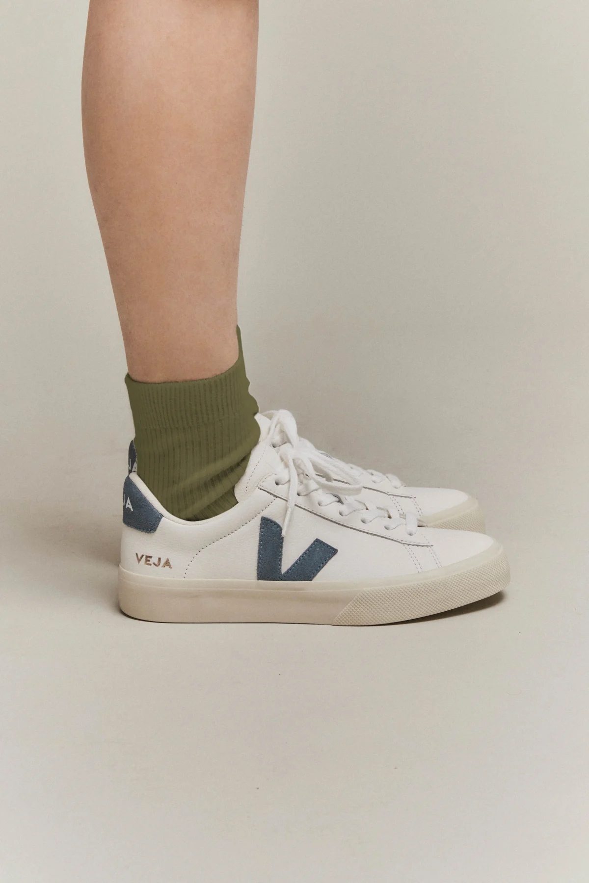 A person wearing olive green socks and white Veja sneakers with blue accents stands on a neutral background. The photo shows a side view of their lower legs and feet.
