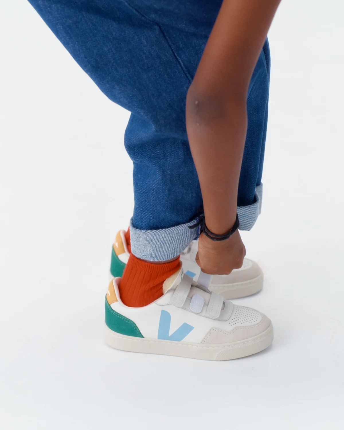 A person wearing blue jeans and orange socks adjusts the cuff of their pants. They are wearing white sneakers with Velcro straps, green and yellow accents, and a light blue V on the side.