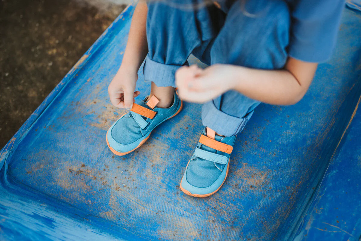 A child wearing blue jeans and blue sneakers with orange straps sits on a blue surface, fastening one shoe. The childs face is not visible in the image.