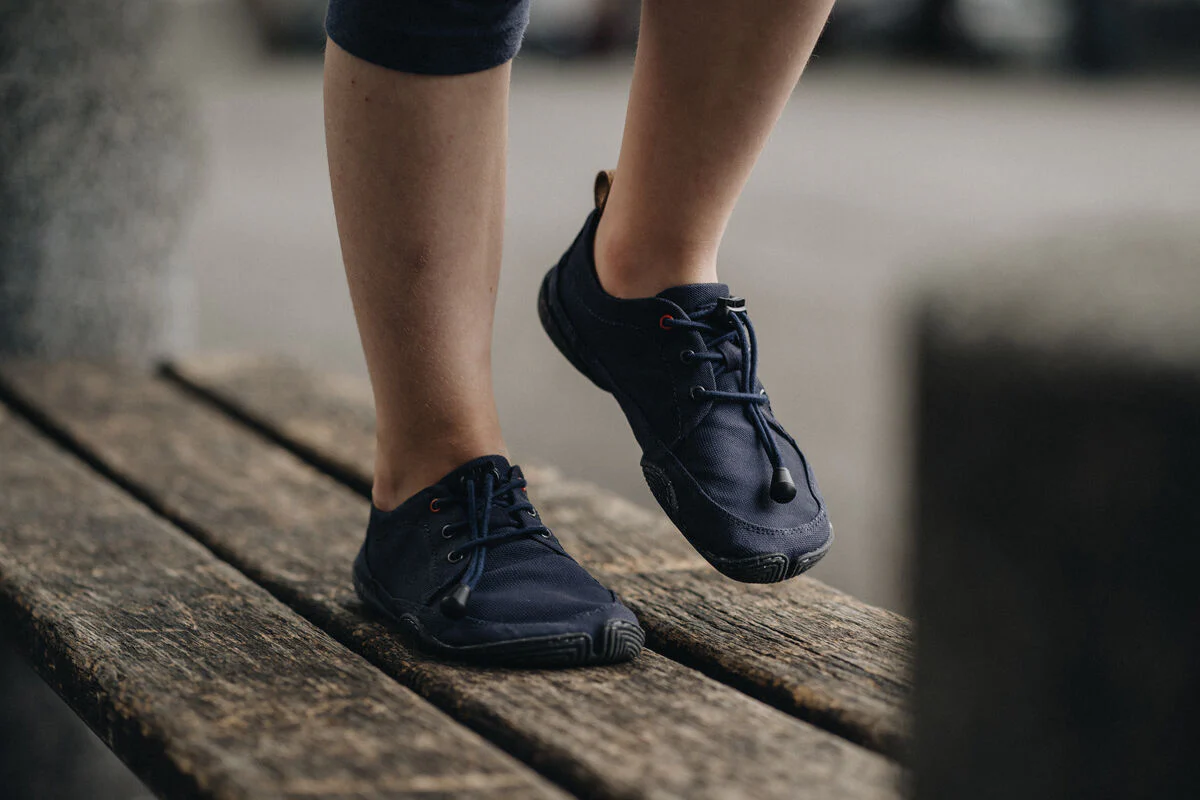 A person wearing dark blue sneakers stands on a weathered wooden bench, with one foot flat and the other raised on its toes. The person is dressed in dark leggings. The background is blurred.