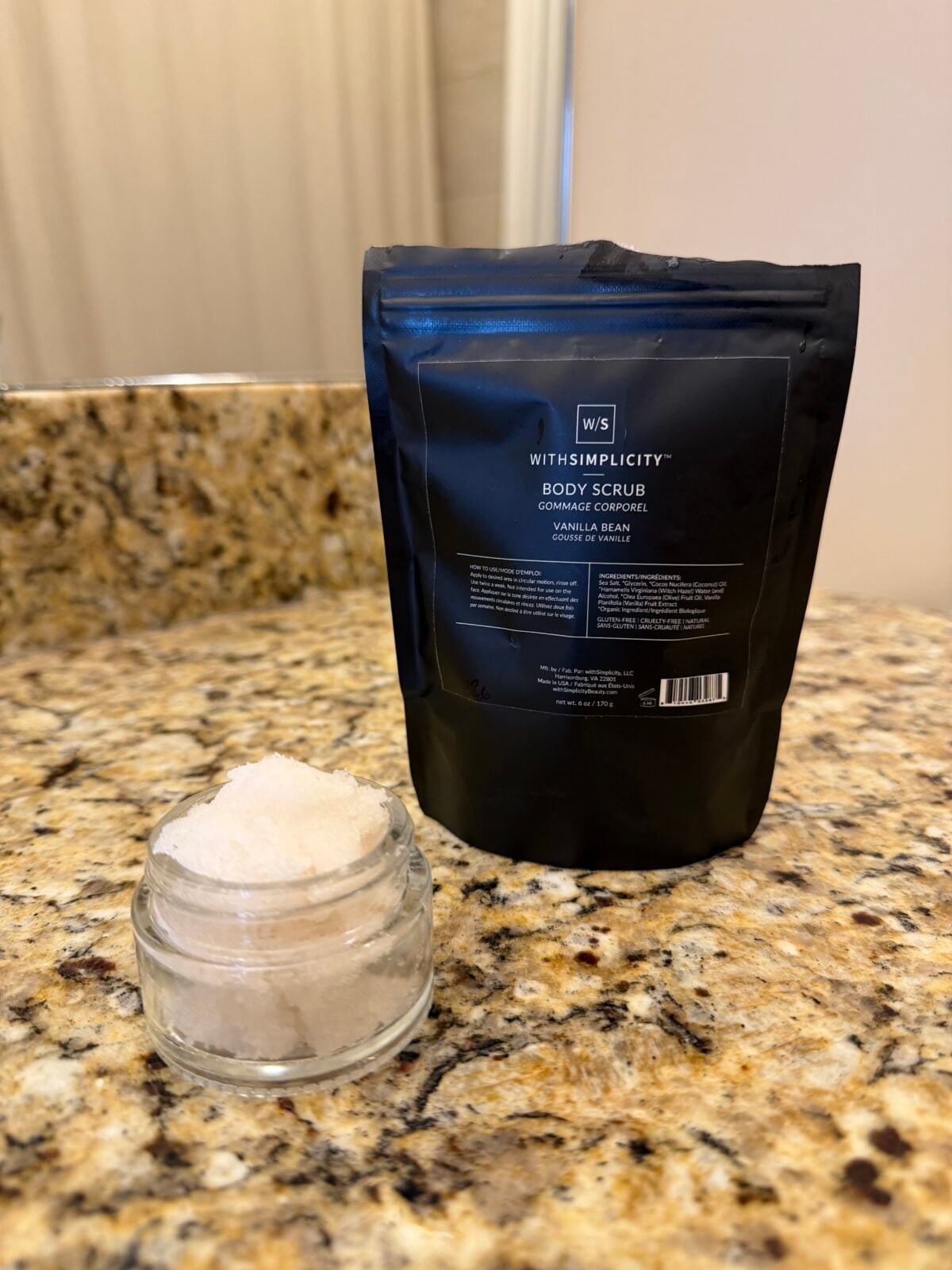 A small glass jar of body scrub sits on a granite countertop in front of a black resealable bag labeled WITHSIMPLICITY Body Scrub Vanilla Bean. A bathroom mirror and curtain are visible in the background.