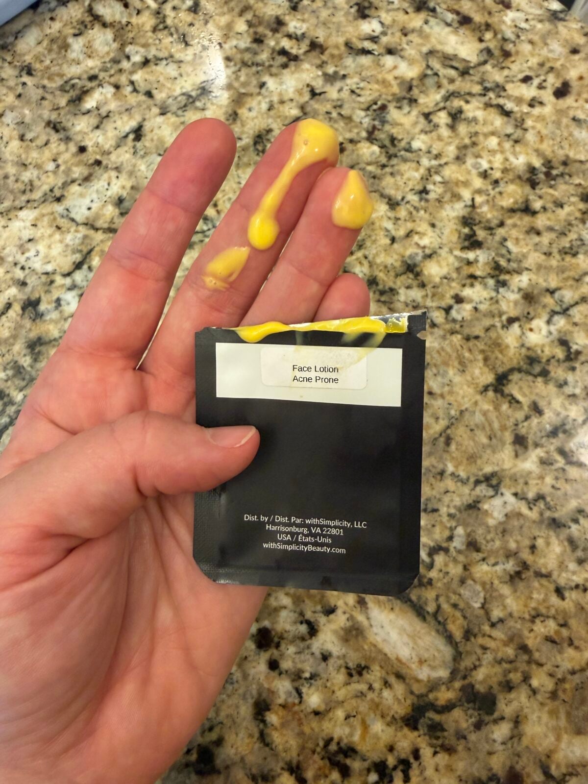 A hand holds an open packet labeled Face Lotion Acne Prone with yellow lotion smeared across the fingers. The background is a granite countertop.