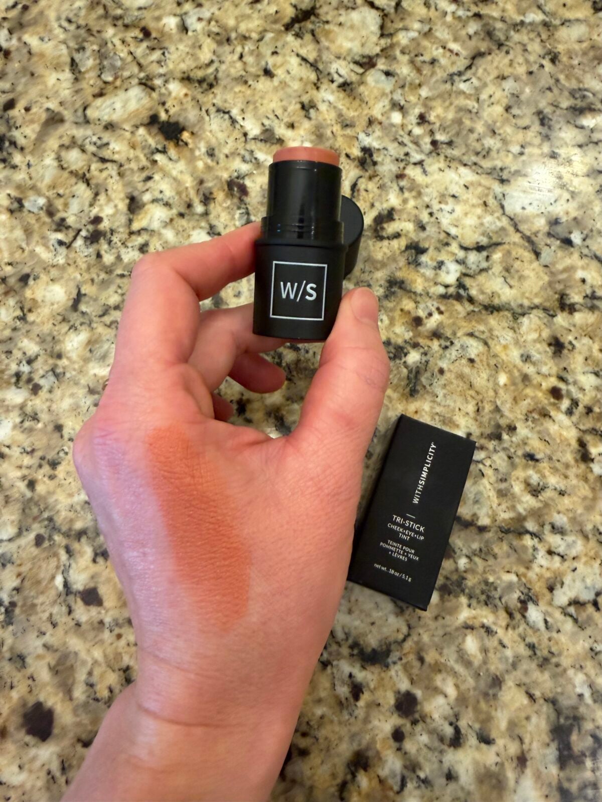 A hand holds a stick of cream makeup labeled W/S against a granite countertop, with a swatch of the product applied on the back of the hand. The makeup sticks cap and box are also visible nearby.