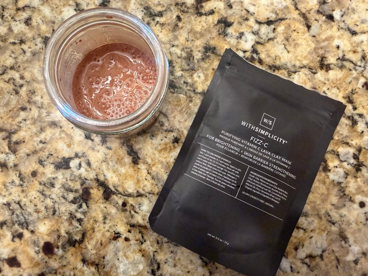 An open glass jar containing a bubbly, pinkish mixture sits on a speckled granite countertop next to a black packet labeled “WITHSIMPLICITY FIZZ C Purifying Pink Lava Clay Mask.”.