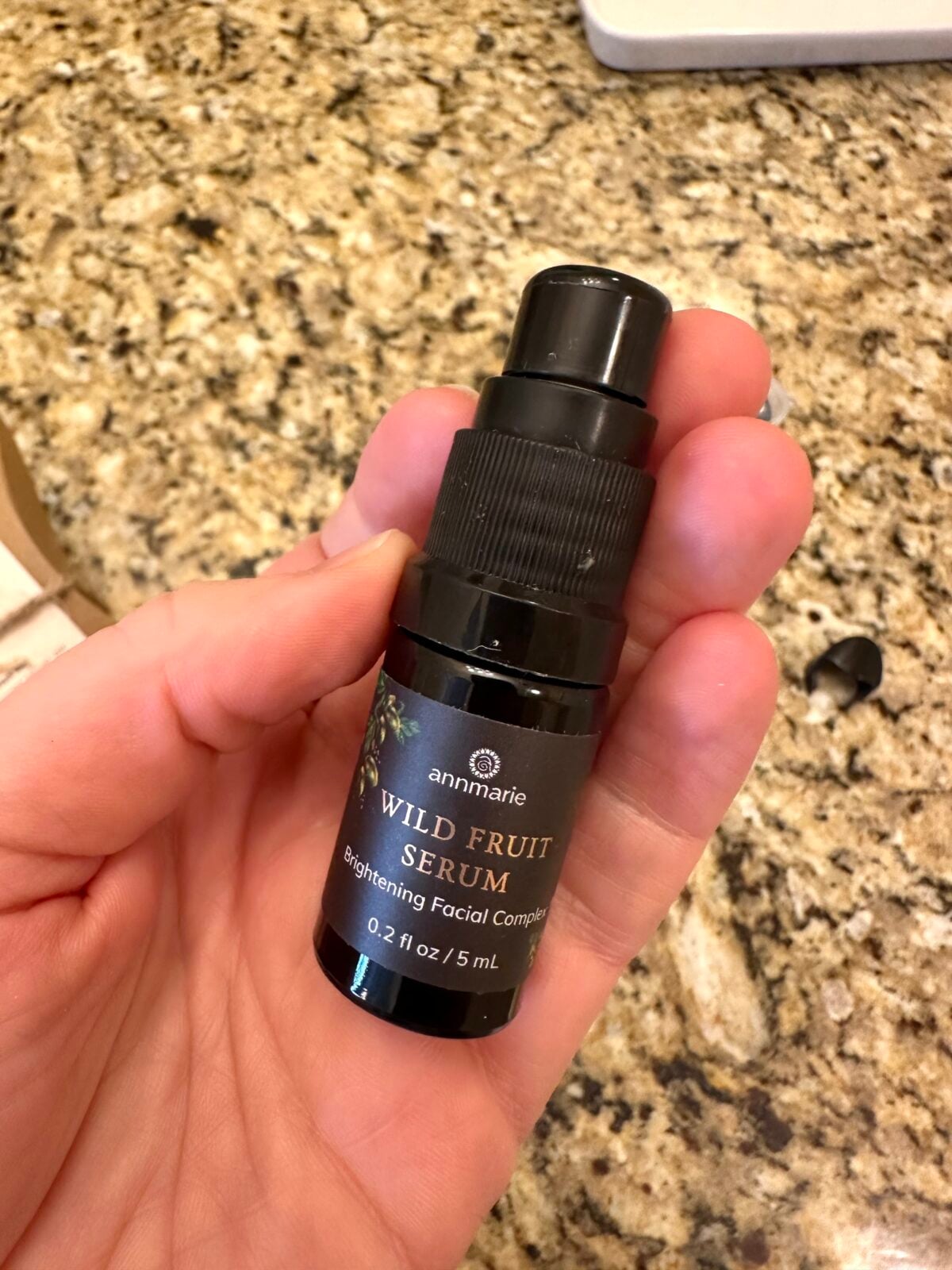 A hand is holding a small 0.2 fl oz (5 mL) black bottle of Annmarie Wild Fruit Serum, labeled as a Brightening Facial Complex, over a granite countertop.