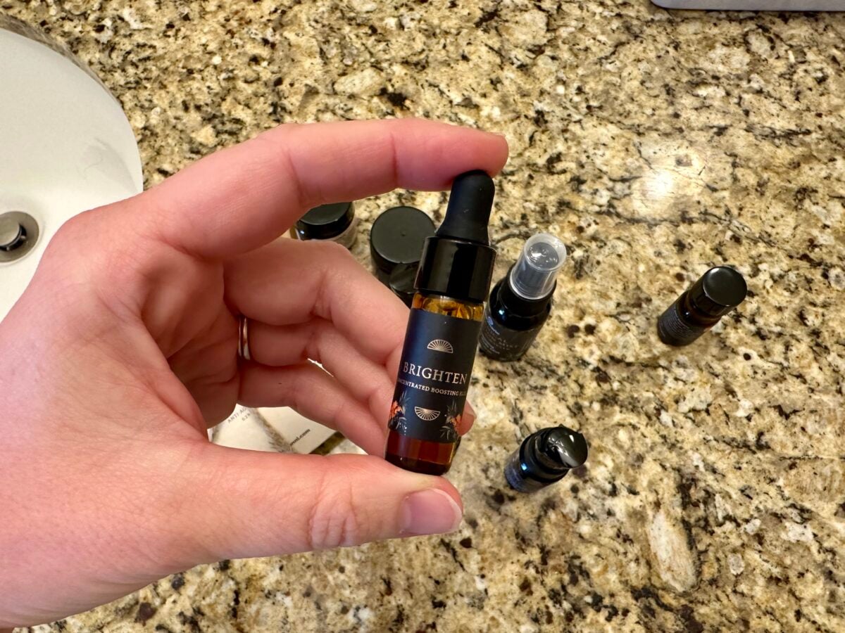 A hand holds a small brown glass dropper bottle labeled BRIGHTEN over a granite countertop with several other small skincare bottles and containers scattered nearby.