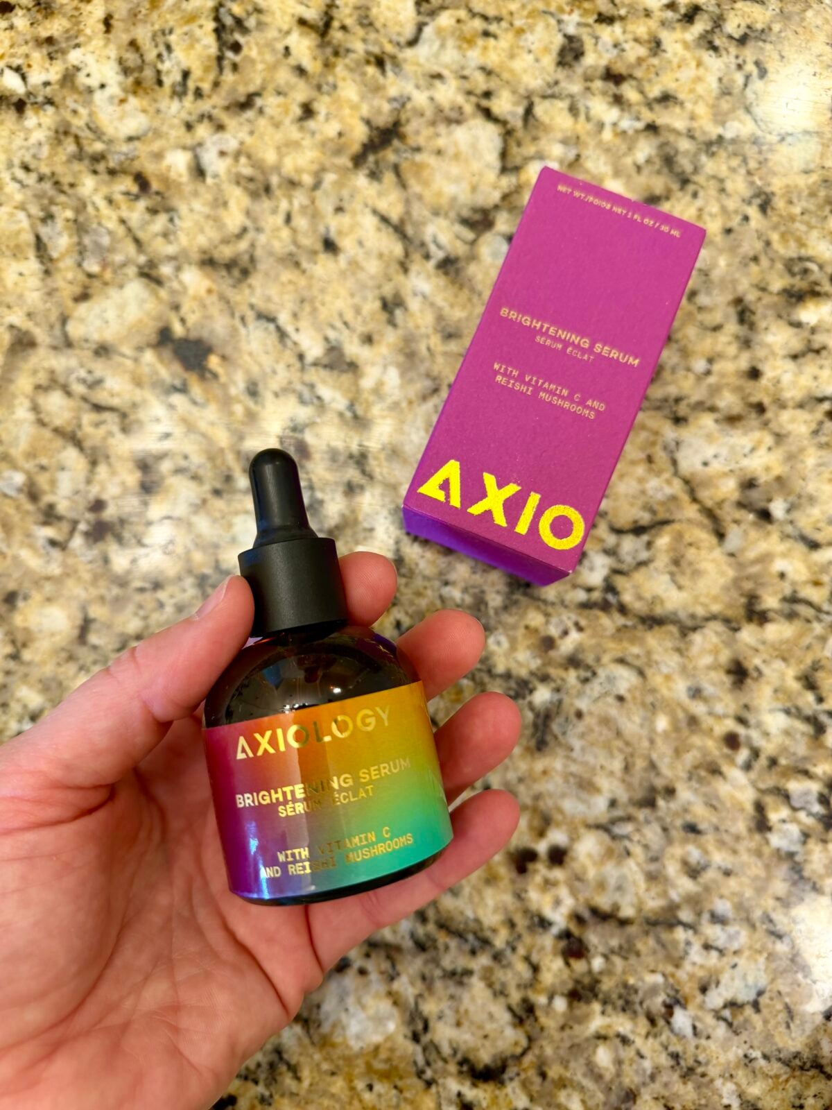 A hand holds a bottle of Axiology Brightening Serum with Vitamin C, Niacinamide, and Mushrooms. A matching purple and yellow product box sits on a granite countertop in the background.