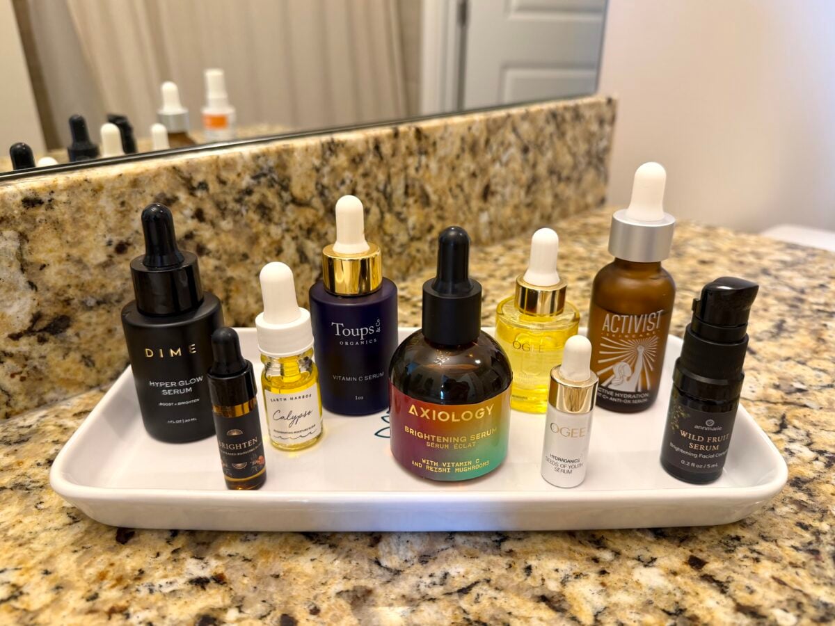 A white tray holds nine assorted skincare serum bottles, including dropper bottles and spray bottles, all arranged neatly on a granite bathroom countertop in front of a mirror.