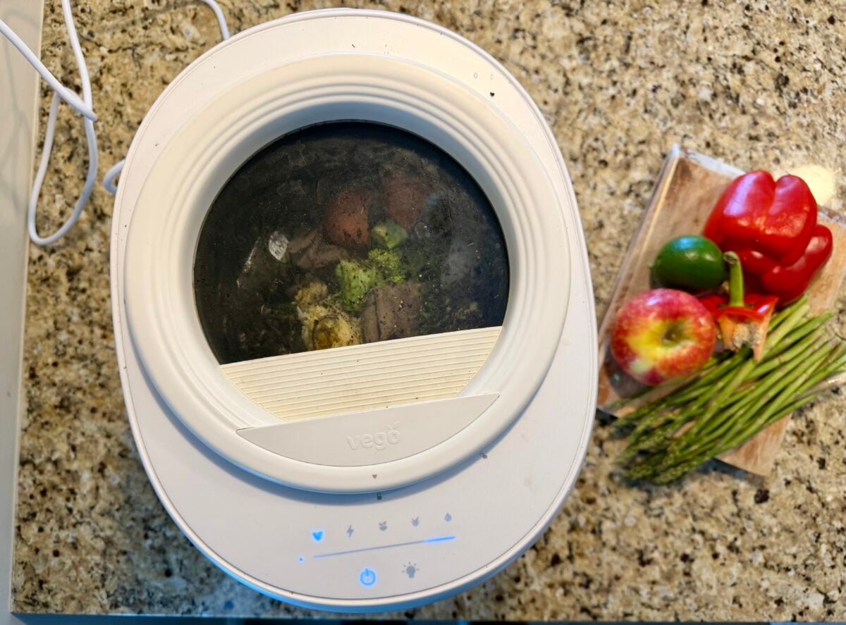 A white countertop food composter with a transparent lid is processing food scraps. Next to it, a cutting board holds a red bell pepper, a lime, an apple, asparagus, and a potato on a speckled granite surface.