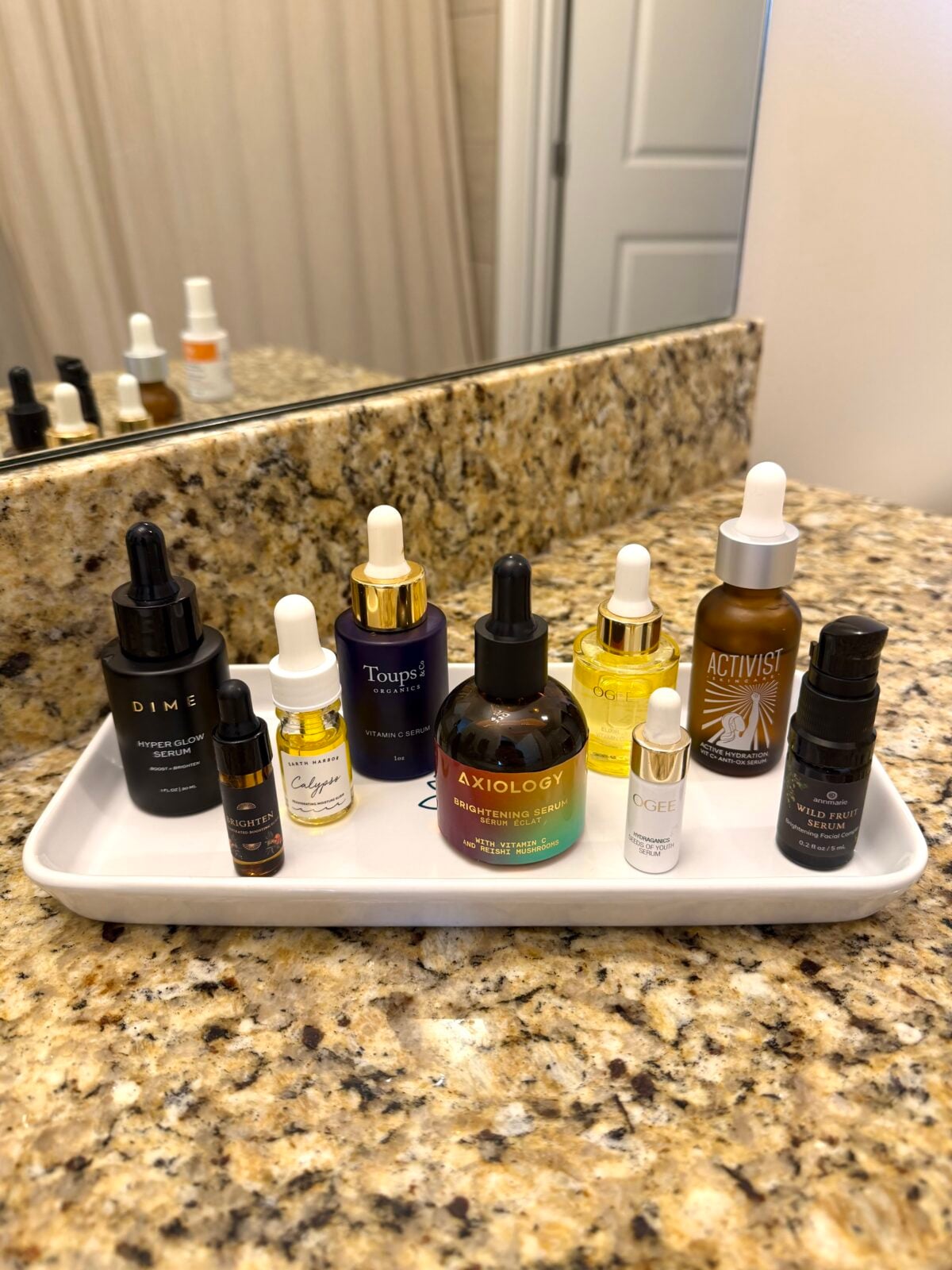 A white tray on a granite bathroom counter holds various skincare bottles and droppers of different shapes, sizes, and colors, arranged neatly in front of a mirror.