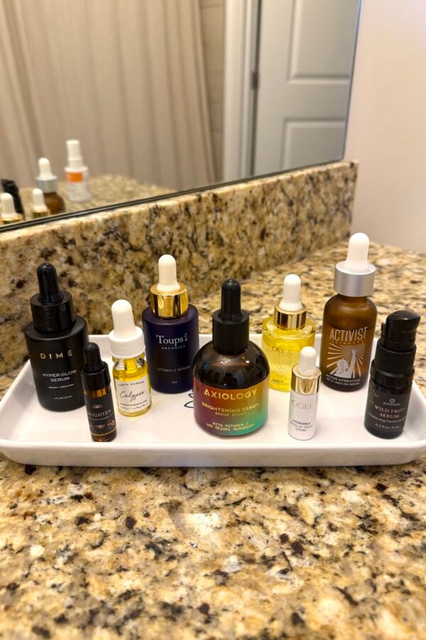 A white tray on a granite bathroom counter holds various skincare bottles and droppers of different shapes, sizes, and colors, arranged neatly in front of a mirror.