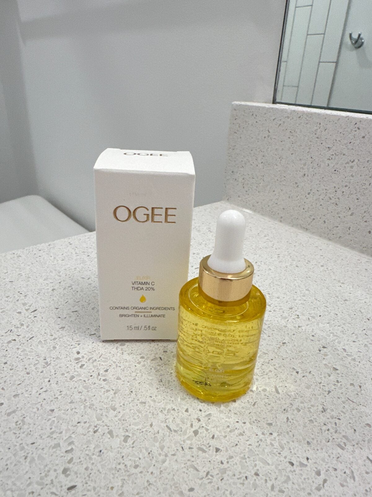 A bottle of OGEE Vitamin C serum with a white and gold dropper cap stands in front of its matching box on a white speckled bathroom countertop, with a mirror and tiled wall in the background.